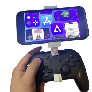 May include: A white phone holder attached to a black Nintendo Switch controller. The phone is displaying a screen with various app icons, including the Apple App Store icon, a game icon, and a magazine icon. The phone holder is attached to the controller with a white clip.