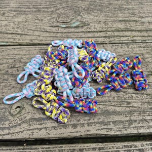 May include: A collection of colorful paracord zipper pulls in various shades of blue, yellow, purple, and rainbow. Each pull is hand-knotted with a loop for easy attachment. The pulls are arranged on a weathered wooden surface.