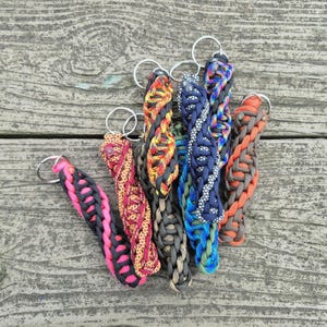 May include: A collection of handmade paracord keychains in various colors, including pink, gray, orange, blue, and brown. Each keychain features a braided design and a silver-colored keyring. The keychains are arranged on a weathered wooden surface.