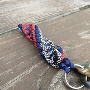 May include: A red, white, and blue paracord keychain with a silver key ring. The keychain is made of braided paracord and has a knot at the end.