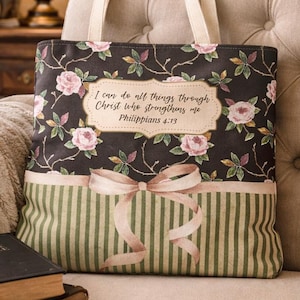 May include: A tote bag featuring a dark floral pattern with pink roses and green leaves, a central cream-colored label with the text 'I can do all things through Christ who strengthens me Philippians 4:13', and a light pink bow over green stripes.
