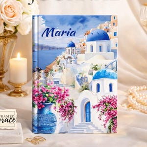 May include: A custom journal with the name "Maria" on the cover. The cover features a watercolor painting of a coastal village with white buildings, blue domes, and vibrant pink flowers. The words "CUSTOM JOURNAL" are at the top.