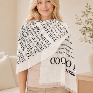 Può includere: Una sciarpa color crema con testo nero con citazioni ispiratrici. La sciarpa ha un bordo sfrangiato ed è drappeggiata sulle spalle. La donna indossa un top rosa chiaro e pantaloni beige.