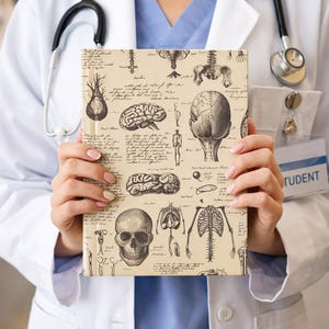 May include: A beige notebook featuring anatomical illustrations of the human brain, skull, and skeleton. The notebook has a vintage aesthetic with handwritten text and detailed drawings. The cover is held by a person wearing a white lab coat.