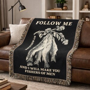 May include: Black throw blanket with a fringed edge. The blanket displays a detailed illustration of fish and botanical elements, with the text "FOLLOW ME" and "AND I WILL MAKE YOU FISHERS OF MEN" in white. The text "MATTHEW 4:19" is also included.