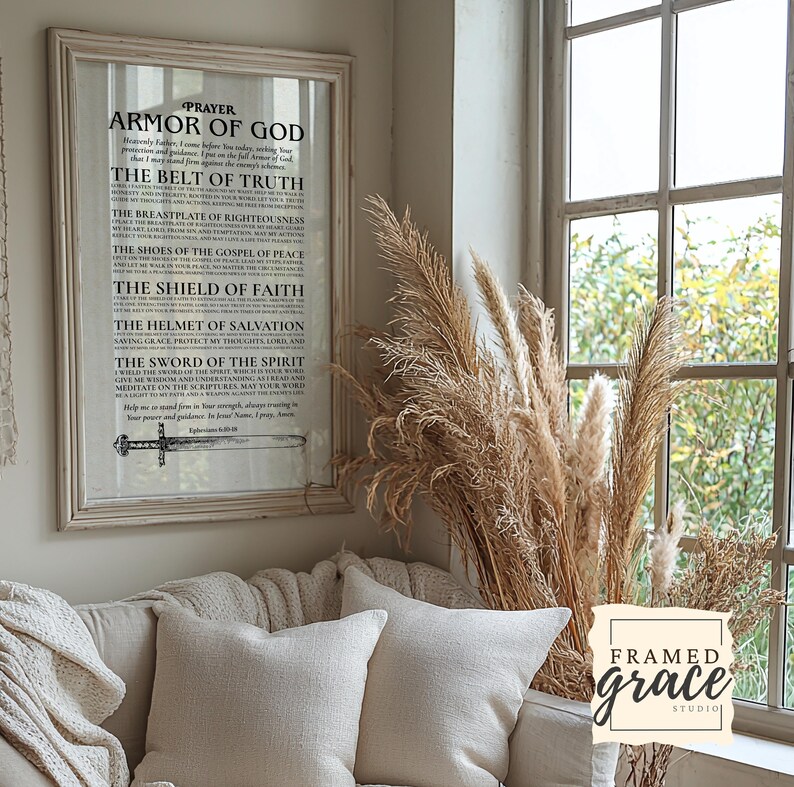 May include: A framed print with the text "Armor of God" in a white frame. The print is displayed above a beige sofa with two white pillows. A window with tall dried grasses is to the right of the print. The print includes the text "Framed Grace Studio".