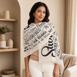May include: A cream-colored scarf with black text. The scarf features phrases like "The Fortress" and "The Branch." The scarf is draped over the shoulders. The scarf has a fringed edge.