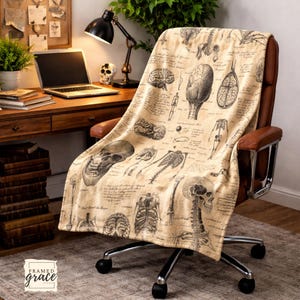 May include: A beige throw blanket draped over a brown office chair. The blanket features black and white anatomical illustrations of the human brain, skull, and skeletal system, with handwritten script. The blanket is on a chair with a silver base.