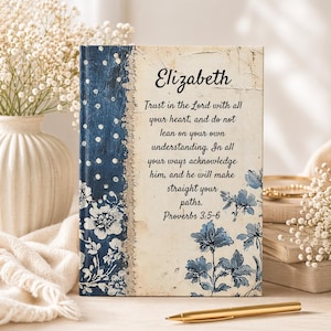 May include: A personalized journal with the name "Elizabeth" on the cover. The journal features a blue floral and polka dot design on the spine and a cream-colored background with a Bible verse. A gold pen rests in front.