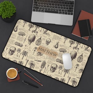 May include: Anatomy-themed desk mat featuring illustrations of the human skeletal and organ systems. The beige mat has a black border and includes the name "MATTHEW" on a brown label. A computer mouse rests on the mat.