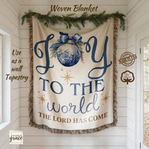 以下が含まれることがあります： 「Joy to the World」の文字と青いクリスマスオーナメントのデザインが施された、クリーム色の織りブランケット。 ブランケットはフリンジ加工が施されており、「Use as a wall tapestry」の文字も確認できます。