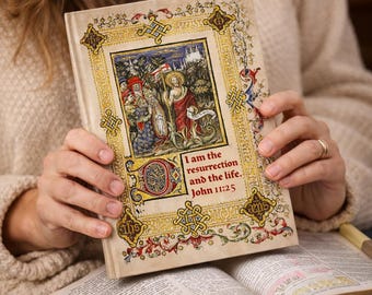 Pode incluir: Um livro de capa dura com uma capa ornamentada e ilustrada apresentando uma cena religiosa e o texto "Eu sou a ressurreição e a vida. João 11:25". O livro é mantido aberto, revelando as páginas de outro livro.