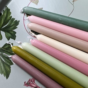 May include: A set of ten taper candles in various shades of green, pink, and brown. The candles are arranged in a fan shape on a light blue surface.