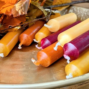 May include: A selection of colourful, cylindrical candles in shades of orange, yellow, and burgundy, arranged on a gold-toned tray. The candles have white wicks and are accompanied by autumn leaves, creating a warm, seasonal display.