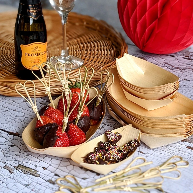 May include: A wooden bowl filled with chocolate-covered strawberries on skewers. The bowl is surrounded by other wooden bowls, some stacked on top of each other. There is a bottle of Prosecco and a glass of wine in the background.