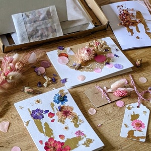 May include: A set of five blank cards with pressed flowers and gold leaf accents. The cards are decorated with pink and white confetti, dried flowers, and a lace doily.