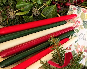 Op de afbeelding: Een verzameling rode, groene en crèmekleurige dinerkaarsen, gerangschikt met feestelijk groen en een decoratief servet. De kaarsen zijn van verschillende lengtes en diagonaal gerangschikt. Een rood fluwelen lint en dennentakjes dragen bij aan het kerstthema.