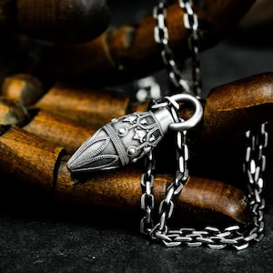 Könnte beinhalten: Eine silberne Halskette mit einem detaillierten, verzierten Anhänger in Form einer Kugel. Der Anhänger hat eine strukturierte Oberfläche und komplizierte Designs. Die Kette ist eine silberne Gliederkette.