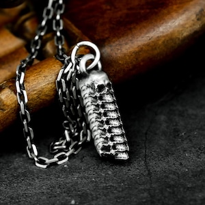 May include: A silver-colored pendant necklace featuring a skull-shaped design. The pendant is a vertical cylinder with multiple skull shapes stacked on top of each other. The chain is a linked design.