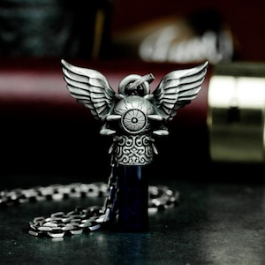 Puede incluir: Un collar con un colgante de plata con un diseño de ojo alado detallado. El ojo está rodeado por una corona y las alas están extendidas. El colgante está unido a una cadena de plata.