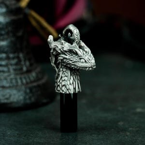 Może przedstawiać: Srebrny korek do butelki w kształcie głowy smoka z misternymi detalami. Smok ma czarną podstawę i stoi na ciemnej powierzchni.