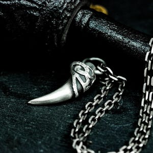 May include: A silver necklace featuring a detailed, textured, fang-shaped pendant. The pendant is attached to a silver chain.