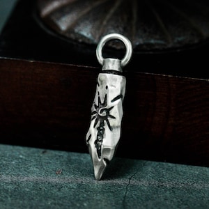 May include: A silver pendant shaped like a jagged rock with a stylised sun design. The pendant has a loop at the top for attaching to a chain.