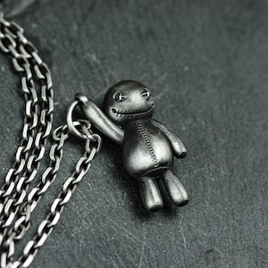 Pode incluir: Um colar de corrente de prata com um pequeno pingente de boneca preta costurado e detalhado. A boneca tem um sorriso e olhos costurados.