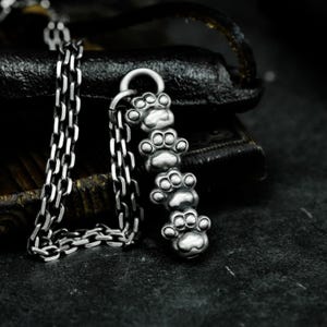 Puede incluir: Un collar de cadena de plata con un colgante que presenta cinco huellas de patas apiladas. El colgante está hecho de plata de ley y tiene un acabado pulido.
