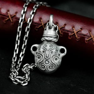 May include: A silver necklace with a detailed, ornate pendant shaped like a small bottle. The pendant has a textured surface with intricate designs.