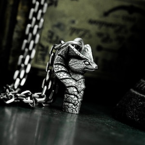 Może przedstawiać: Srebrny naszyjnik z wisiorkiem w kształcie głowy kobry. Głowa węża jest wyrzeźbiona z teksturowanymi łuskami i skupionym spojrzeniem. Łańcuszek jest w klasycznym stylu. Tło jest ciemne.