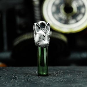以下が含まれることがあります： 閉じた目と額に花柄のデザインが施された、精巧な狐の頭部が描かれたシルバーチャーム。チャームは緑色のガラス管に取り付けられています。