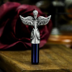 May include: A silver bottle stopper with an ornate angel design. The angel has outstretched wings and a detailed face. The stopper is attached to a blue glass tube.