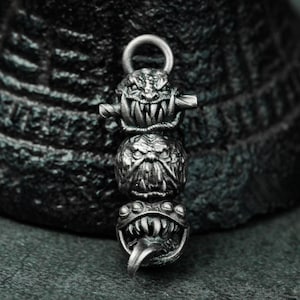 May include: A silver charm necklace featuring three detailed, grotesque monster faces. The top face has a wide open mouth with sharp teeth, the middle face has a closed mouth with a furrowed brow, and the bottom face has a wide open mouth with a long, pointed tongue.