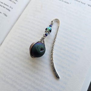 May include: A silver bookmark with a decorative edge and a dark green iridescent gemstone wrapped in silver wire. The gemstone has a small hole in the center. The bookmark is adorned with small blue and green beads.