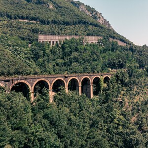 Puede incluir: Un puente de arco de piedra cruza un valle, con exuberantes árboles verdes y follaje que cubren las laderas de las colinas circundantes.