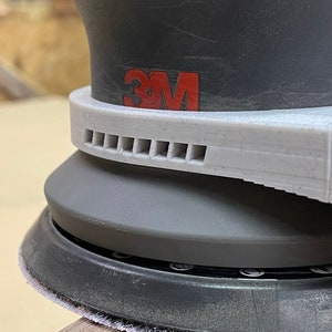 May include: Close-up of a gray and black 3M random orbital sander with a white dust collection ring and a vent with multiple holes.