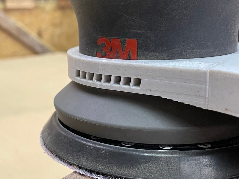 May include: Close-up of a gray and black 3M random orbital sander with a white dust collection ring and a vent with multiple holes.