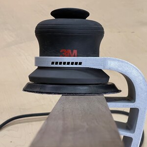 May include: A close-up of a 3M random orbital sander with a white plastic handle attached to a wooden surface. The sander has a black rubber base and a gray plastic ring around the base.