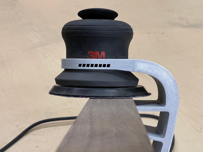 May include: A close-up of a 3M random orbital sander with a white plastic handle attached to a wooden surface. The sander has a black rubber base and a gray plastic ring around the base.