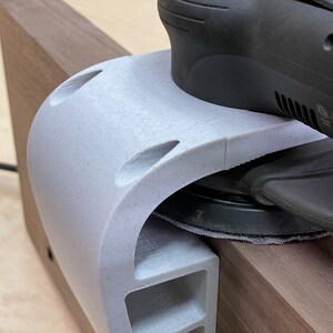 May include: A close-up of a white 3D printed sanding jig being used with a black random orbital sander. The sander is resting on a piece of wood.