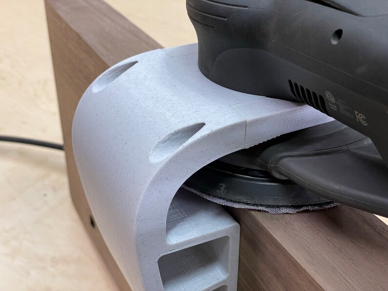 May include: A close-up of a white 3D printed sanding jig being used with a black random orbital sander. The sander is resting on a piece of wood.