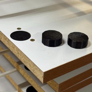 Puede incluir: Primer plano de un componente de armario blanco y color madera con un gran agujero circular y dos objetos cilíndricos negros texturizados. El armario está hecho de paneles en capas.