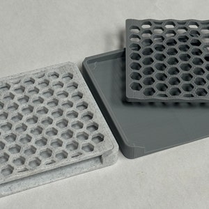 May include: Two square-shaped trays with a honeycomb pattern. One tray is gray and the other is a light gray with a speckled finish. The trays are stacked on top of each other.