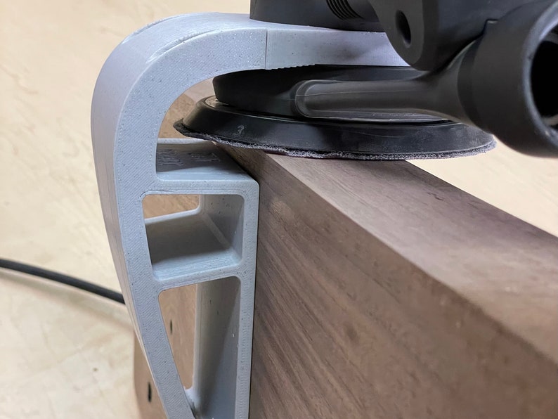 May include: A gray 3D printed sanding jig for woodworking projects. The jig holds a sander in place to help create smooth, even surfaces.