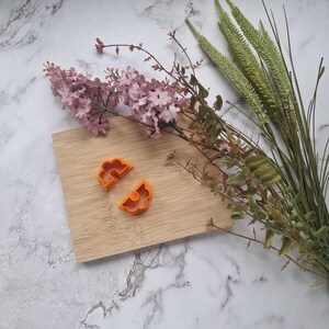 Peut inclure: Deux emportes-pièces en plastique orange en forme de demi-cercles, sur une surface en bois. Les emportes-pièces sont disposés de manière à ressembler à une paire d'yeux.