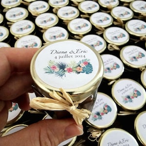 Peut inclure: Plusieurs petits pots en verre avec des couvercles dorés, chacun orné d'une étiquette avec des motifs floraux et le texte "Diana & Eric 4 juillet 2024". Un ruban de raphia est noué autour d'un pot. Les pots sont sur une surface sombre.