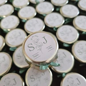 Peut inclure: Petits pots en verre avec des couvercles dorés, chacun orné d'une étiquette circulaire avec les initiales "S" et "J", un motif floral et les noms "Sophie" et "Julian". Les couvercles sont attachés avec un ruban vert clair.