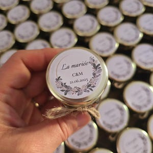 Peut inclure: Petits pots en verre avec des couvercles dorés, attachés avec de la ficelle. Les étiquettes indiquent "La mariée" et "C&M 21.06.2025" entourées d'une couronne florale. Les pots sont probablement des cadeaux de mariage.
