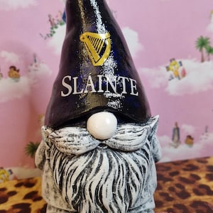 Może przedstawiać: Ceramiczny krasnal ogrodowy z czarną czapką z logo Guinness i napisem "Slainte". Krasnal ma białą brodę i biały nos. Tło jest różowe i w panterkę.